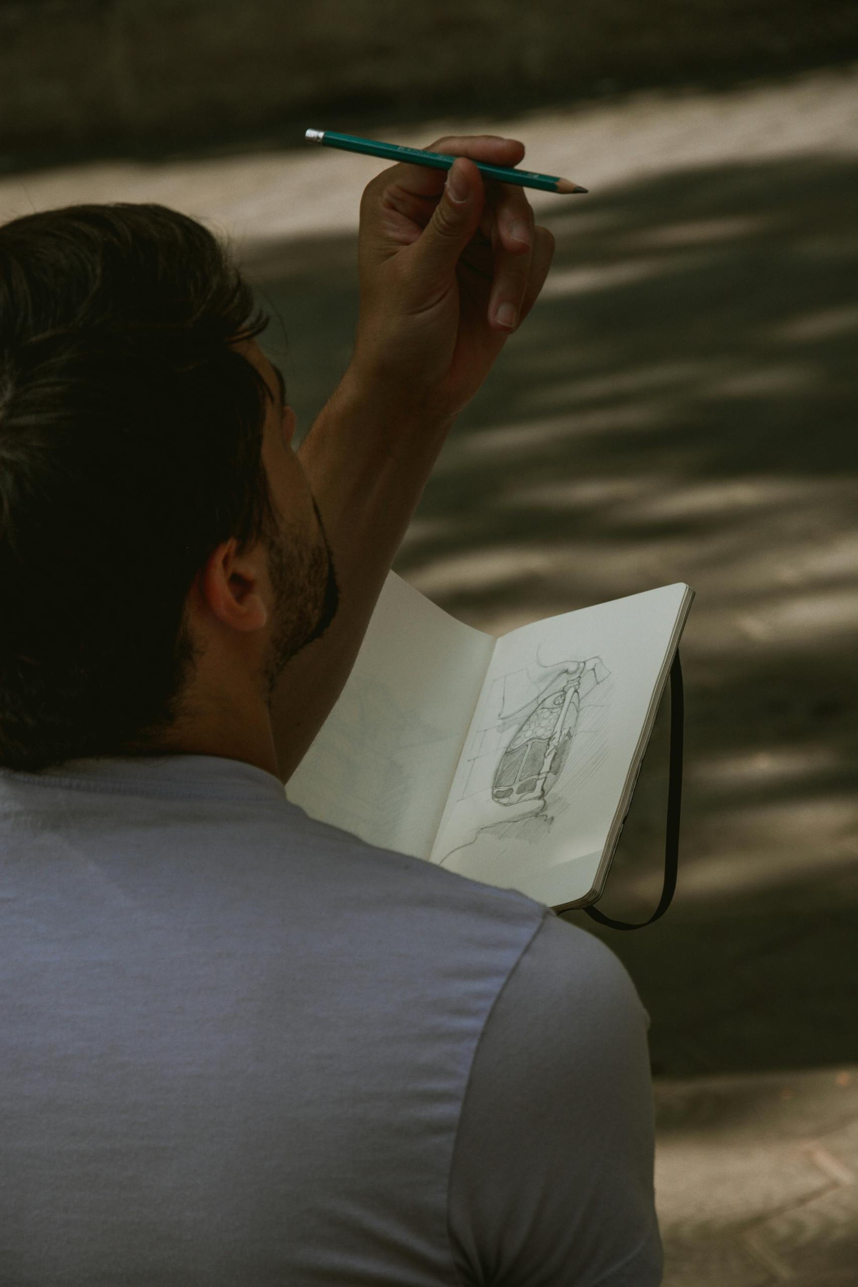 A man sketching a design in a notebook while sitting outdoors, captured in natural light.