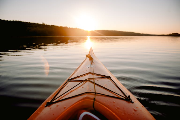 Orange kayak floating on rippling surface of calm river against forest with trees in sundown time in nature on summer evening