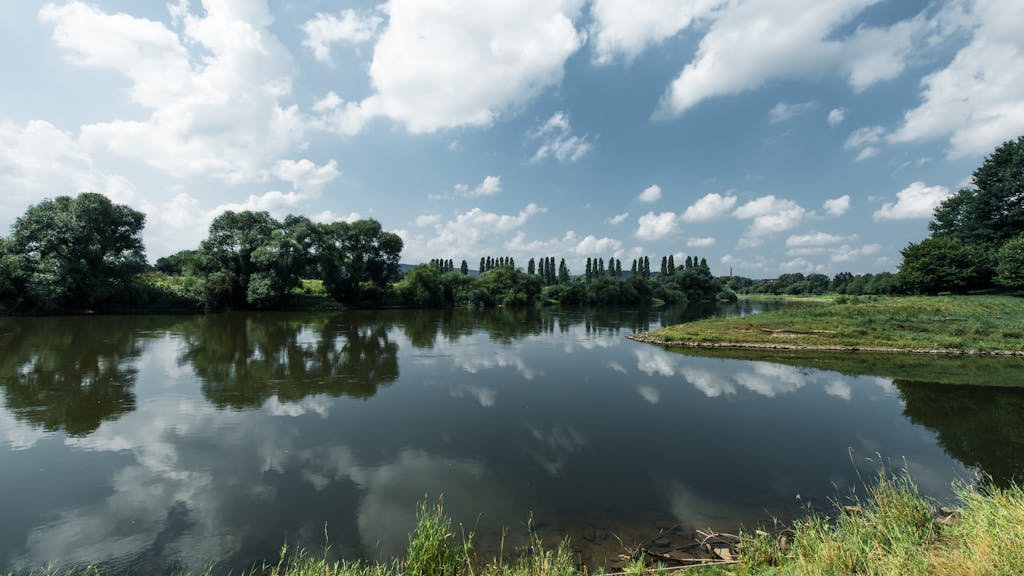Serene river landscape with mirrored trees and cloudy sky in Höxter, Germany.