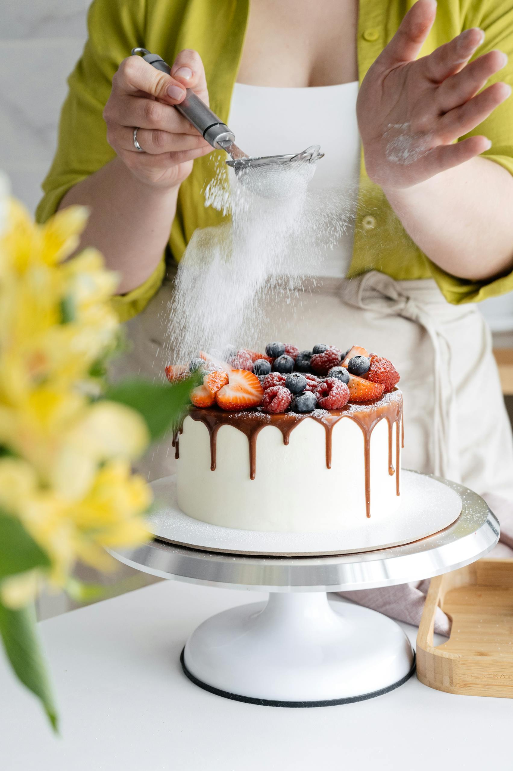 A beautifully decorated cake with fresh berries and icing sugar being dusted on top.