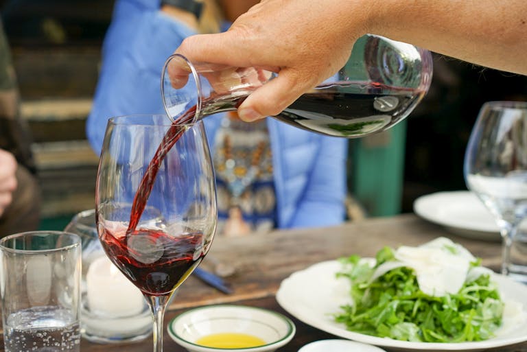A hand pours red wine into a glass at a luxurious dining setting with salad on a wooden table.