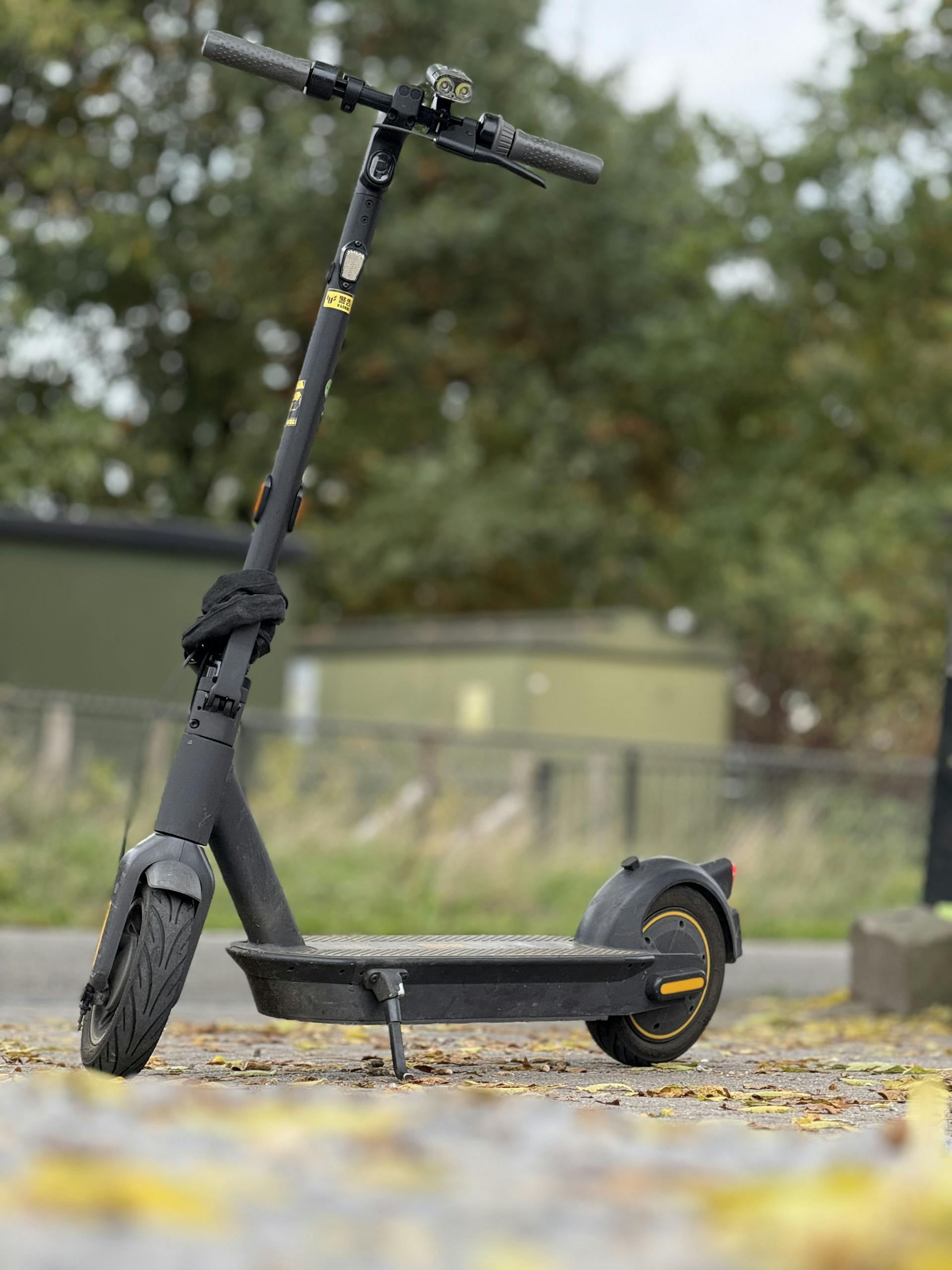 A sleek black electric scooter parked outdoors on a path covered with autumn leaves.