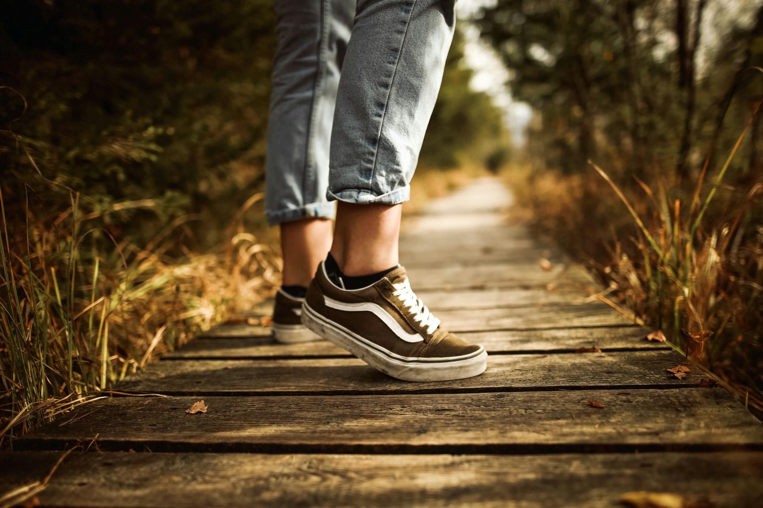 Close-up of a person walking on a wooden path in a park wearing jeans and sneakers during autumn.