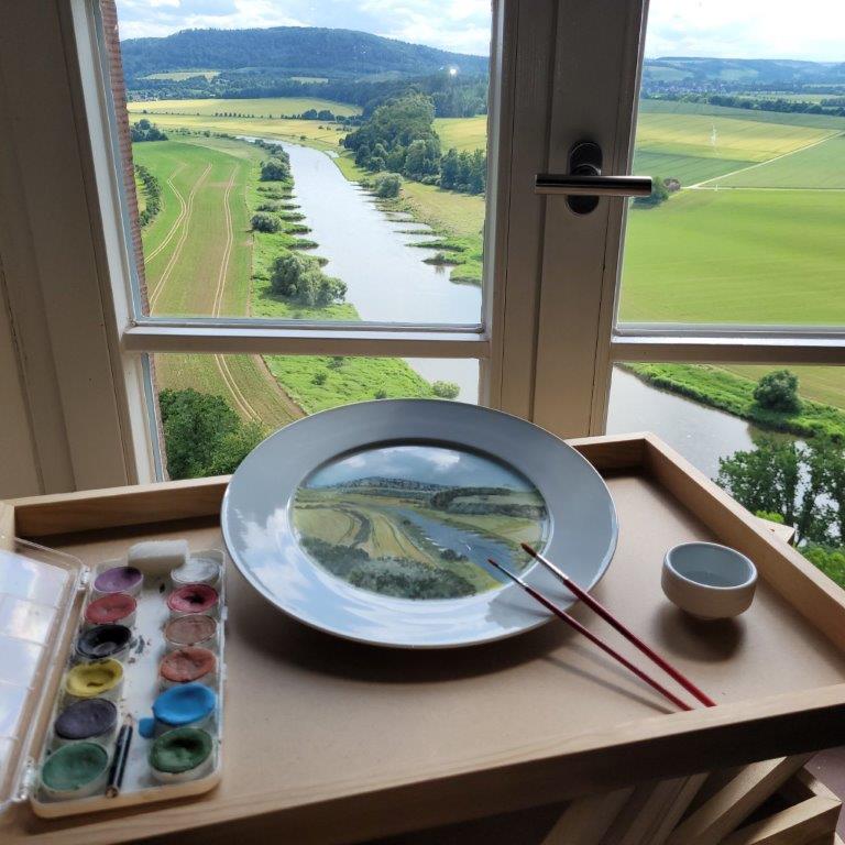 Ein Tablett auf einer Fensterbank mit Blick auf eine grüne Flusslandschaft. Darauf liegen ein bemalter Porzellanteller mit Landschaftsmotiv, zwei feine Pinsel, ein kleiner Wasserbecher und eine Farbpalette. Im Hintergrund schlängelt sich ein Fluss durch Felder und Hügel unter leicht bewölktem Himmel.