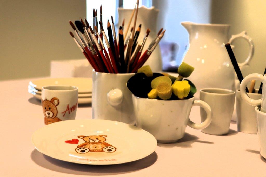 Materialien zum Porzellanmalen mit Pinseln, Schwämmen und weißem Geschirr. Im Vordergrund sind Teller und Tasse mit Teddybär-Motiv zu sehen.
