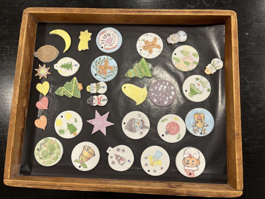 Auf einem Holztablett liegen zahlreiche handbemalte Porzellanhänger in verschiedenen Formen mit winterlichen und weihnachtlichen Motiven wie Tannenbäumen, Schneemännern, Sternen und Herzen.