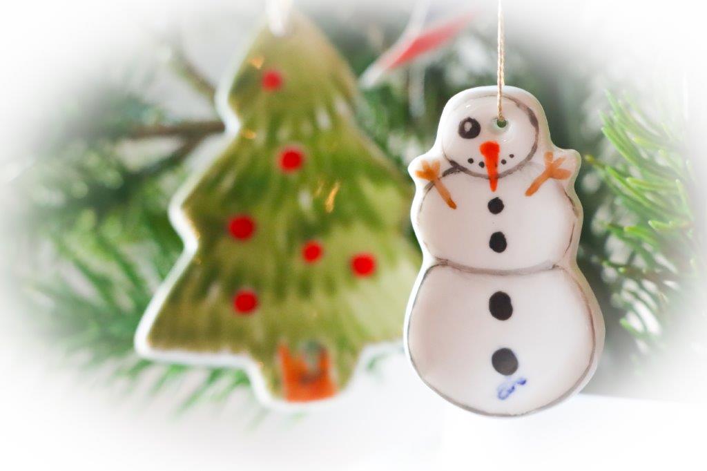 Nahaufnahme von zwei handbemalten Porzellanhängern in Form eines Schneemanns und eines Weihnachtsbaums, die zwischen grünen Tannenzweigen hängen.